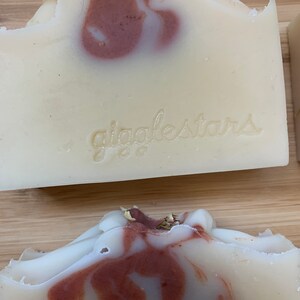 Może przedstawiać: Zbliżenie na ręcznie robione kostki mydła z kremowo-białą bazą i wirującymi bordowymi akcentami. Mydło ma teksturowaną powierzchnię, a na przodzie wytłoczono napis "giggletars". Mydło leży na drewnianej powierzchni.