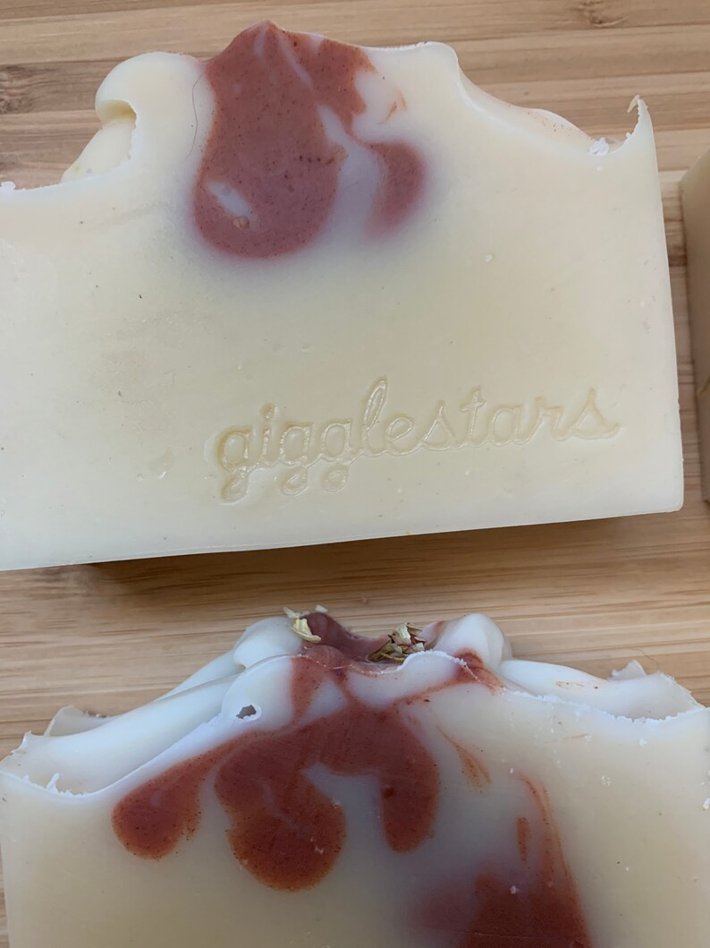 Może przedstawiać: Zbliżenie na ręcznie robione kostki mydła z kremowo-białą bazą i wirującymi bordowymi akcentami. Mydło ma teksturowaną powierzchnię, a na przodzie wytłoczono napis "giggletars". Mydło leży na drewnianej powierzchni.