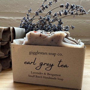 May include: A bar of handmade soap with a brown paper wrapper. The soap is labeled "gigglestars soap co. earl grey tea Lavender & Bergamot Small Batch Handmade Soap". Dried lavender flowers are on top of the soap.