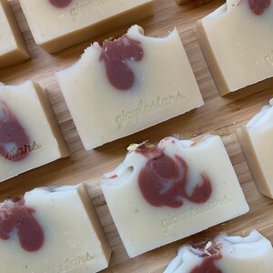 Może przedstawiać: Zbliżenie na ręcznie robione kostki mydła z kremowo-białą bazą i bordowym wzorem w wiry. Kostki są opatrzone napisem "gigglestars". Niekt&oacute;re kostki mają odgryziony kawałek, a niekt&oacute;re mają złote płatki.