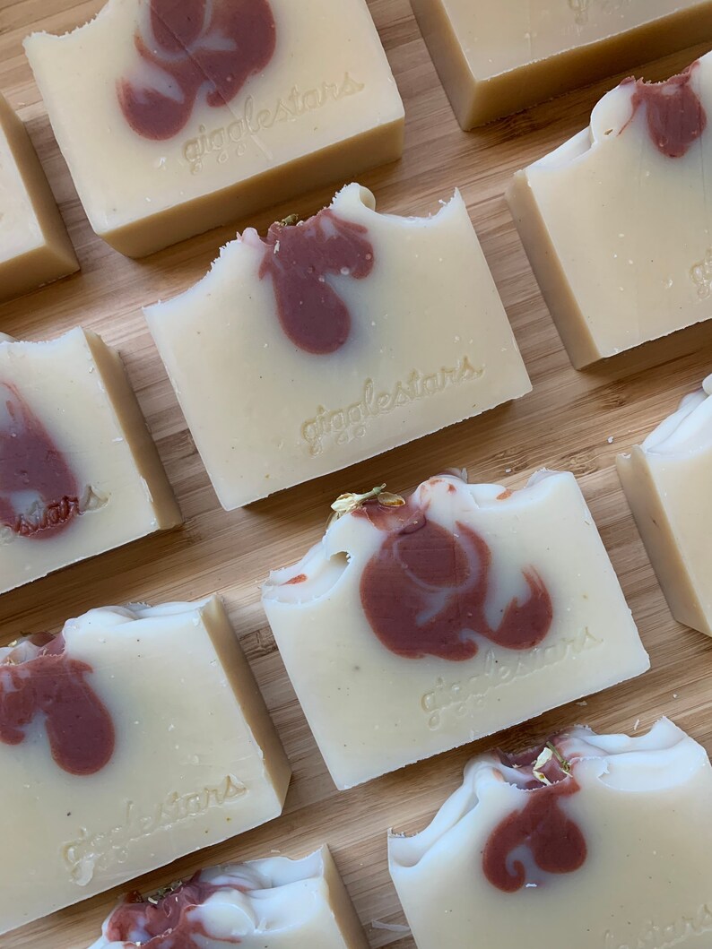 Może przedstawiać: Zbliżenie na ręcznie robione kostki mydła z kremowo-białą bazą i bordowym wzorem w wiry. Kostki są opatrzone napisem "gigglestars". Niekt&oacute;re kostki mają odgryziony kawałek, a niekt&oacute;re mają złote płatki.