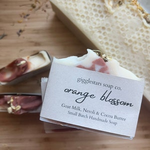 Może przedstawiać: Zbliżenie na ręcznie robione kostki mydła. Jedna kostka jest oznaczona jako "orange blossom" z wymienionymi składnikami. Mydło ma biało-r&oacute;żowy marmurkowy wz&oacute;r. Inna kostka ma wz&oacute;r plastra miodu. Mydła leżą na drewnianej powierzchni.