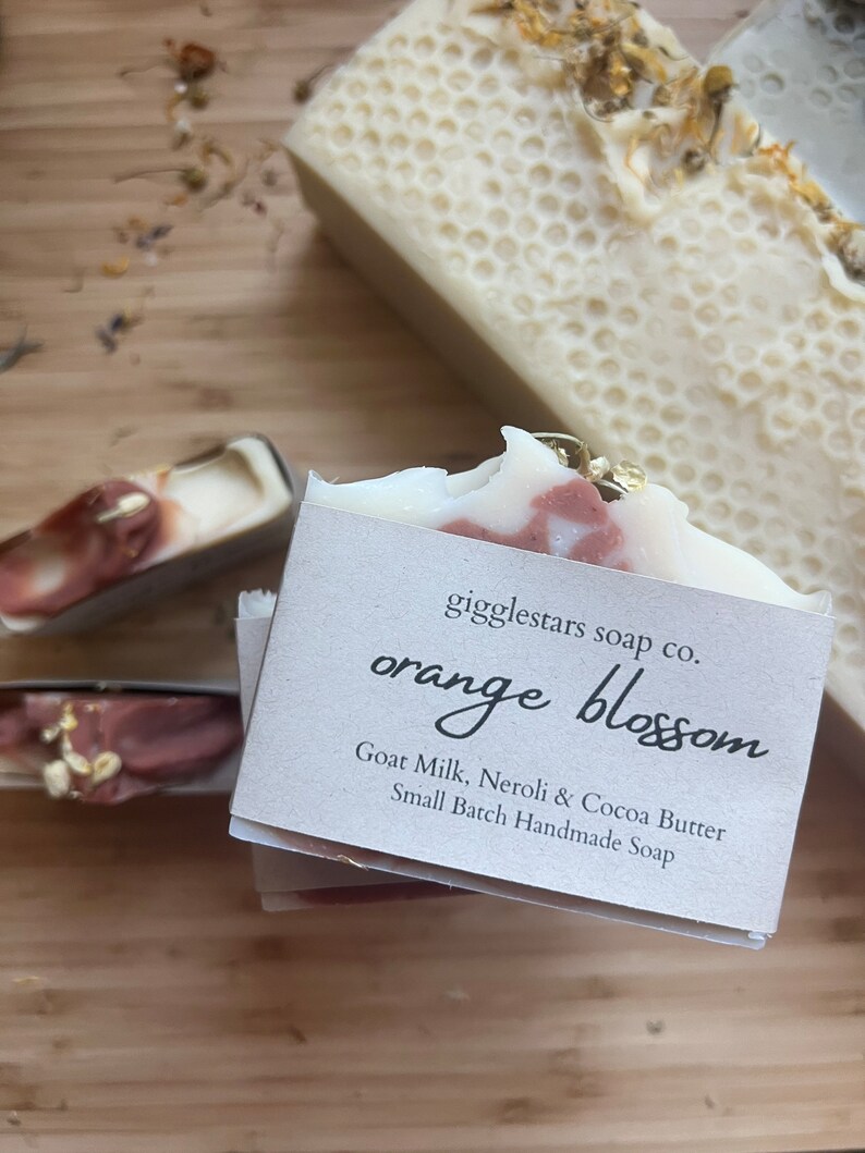 Może przedstawiać: Zbliżenie na ręcznie robione kostki mydła. Jedna kostka jest oznaczona jako "orange blossom" z wymienionymi składnikami. Mydło ma biało-r&oacute;żowy marmurkowy wz&oacute;r. Inna kostka ma wz&oacute;r plastra miodu. Mydła leżą na drewnianej powierzchni.