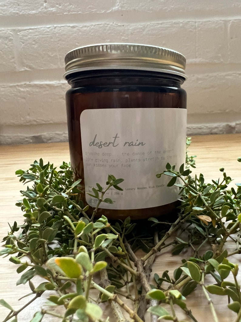 May include: A brown glass candle jar with a silver lid. The label reads "desert rain" and has text about the scent. The candle is sitting on a bed of green foliage.