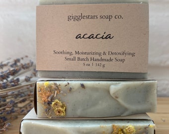 Peut inclure: Trois savons artisanaux à l'acacia, chacun avec une base gris clair et des flocons de fleurs séchées. La barre supérieure porte une étiquette en papier brun sur laquelle est inscrit "gigglestars soap co. acacia Soothing, Moisturizing & Detoxifying Small Batch Handmade Soap 5 oz | 142 g"