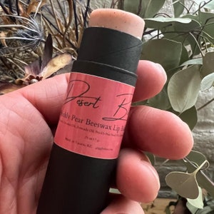 May include: A black tube of lip balm with a pink label that says "Desert Bloom Prickly Pear Beeswax Lip Balm". The tube is made of cardboard and has a black cap. The lip balm is a light pink color.