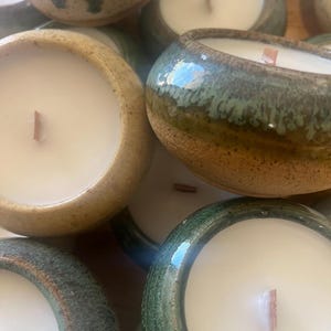 May include: Close-up of several ceramic candles with white wax and wooden wicks. The ceramic bowls have a rustic, glazed finish in shades of green, brown, and tan. The candles are arranged closely together.