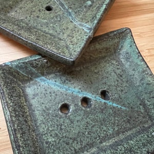 May include: Two green ceramic soap dishes with three drainage holes each. The dishes are textured and have a speckled appearance.