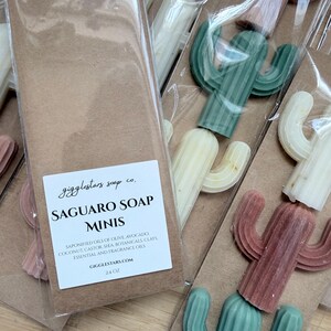 May include: Multiple cactus-shaped soap bars in various colors including green, white, and terracotta, packaged in clear plastic sleeves on brown cardstock. The label reads GIGGLESTARS SOAP CO. SAGUARO SOAP MINIS, 2.4 OZ, and lists ingredients.