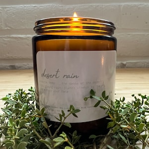 May include: A brown glass candle jar with a white label that reads "desert rain". The label has text about the scent and the candle is lit with a flame.