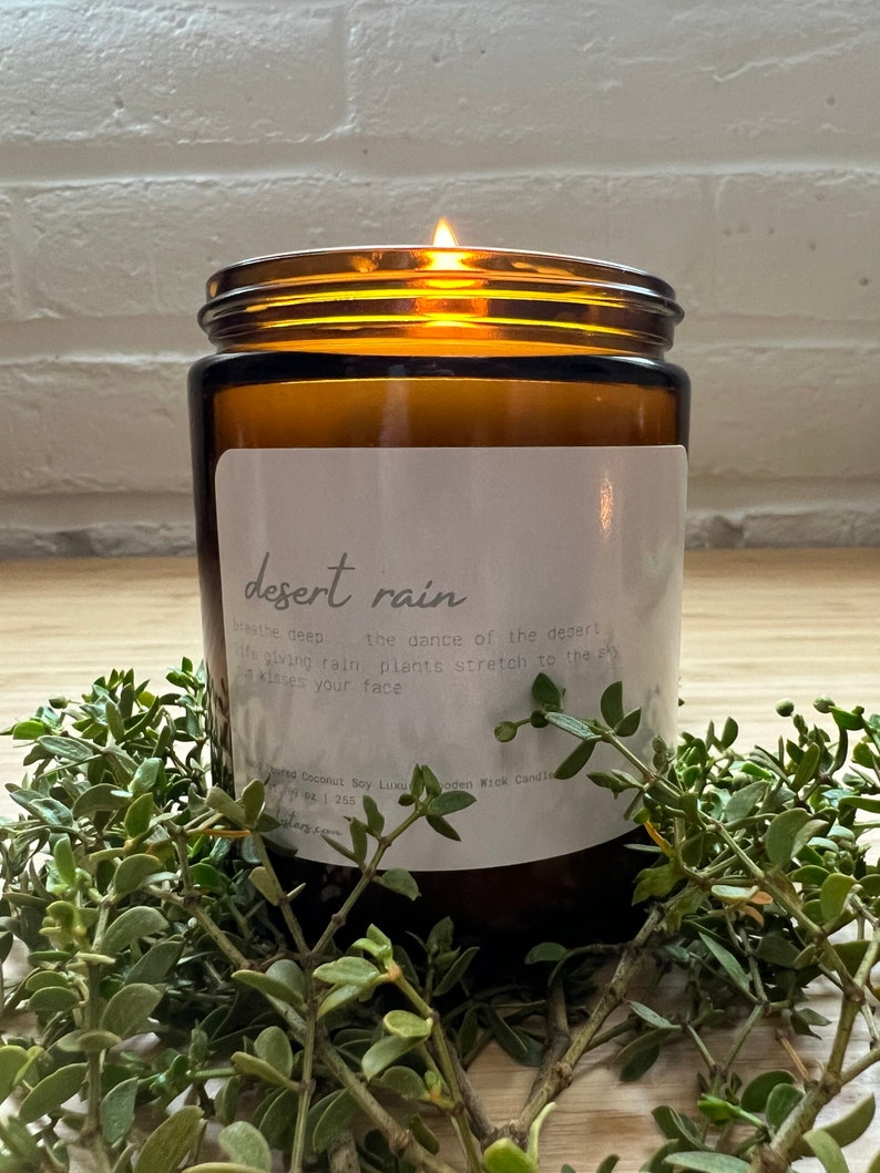 May include: A brown glass candle jar with a white label that reads "desert rain". The label has text about the scent and the candle is lit with a flame.