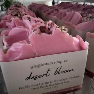 May include: A bar of pink soap topped with dried botanical pieces, wrapped in a white paper label that reads gigglestars soap co. desert bloom Prickly Pear Cactus & Himalayn Rhubarb Small Batch Handmade Soap 5 oz | 142 g, with more bars behind.