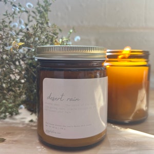May include: A brown glass candle jar with a white label that reads "desert rain" and a description of the scent. The candle is sitting on a wooden surface.