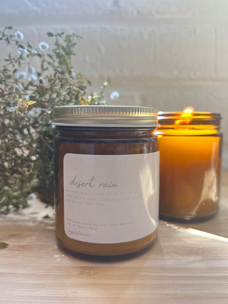May include: A brown glass candle jar with a white label that reads "desert rain" and a description of the scent. The candle is sitting on a wooden surface.