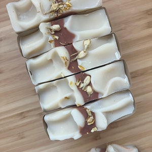 Może przedstawiać: Pięć kostek mydła ręcznie robionego z białym i brązowym wzorem w wiry, zwieńczonych suszonymi kwiatami. Mydło jest zapakowane w brązową papierową osłonę. Na opakowaniu widoczny jest napis "giggletars soap co. sage blossom".