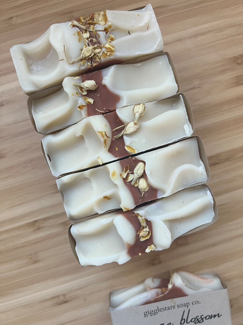 Może przedstawiać: Pięć kostek mydła ręcznie robionego z białym i brązowym wzorem w wiry, zwieńczonych suszonymi kwiatami. Mydło jest zapakowane w brązową papierową osłonę. Na opakowaniu widoczny jest napis "giggletars soap co. sage blossom".