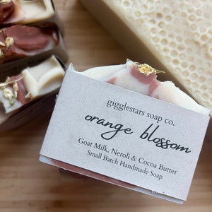 Może przedstawiać: Zbliżenie na ręcznie robione kostki mydła. Jedna kostka jest oznaczona jako "orange blossom" z wymienionymi składnikami. Mydło ma biało-r&oacute;żowy wz&oacute;r z wirami i złotymi akcentami. Kolejna kostka ma wz&oacute;r plastra miodu. Na drewnianej powierzchni.