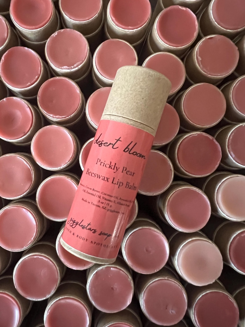 May include: A close-up of a brown cardboard tube with a pink lip balm inside. The tube is labeled "desert bloom prickly pear beeswax lip balm". The lip balm is surrounded by many other similar tubes.