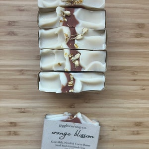 Może przedstawiać: Pięć ręcznie robionych mydeł z białym i brązowym wzorem w wiry, posypanych suszonymi kwiatami. Mydło jest oznaczone "orange blossom" i wykonane z mleka koziego, neroli i masła kakaowego. Każdy kawałek waży 142g.