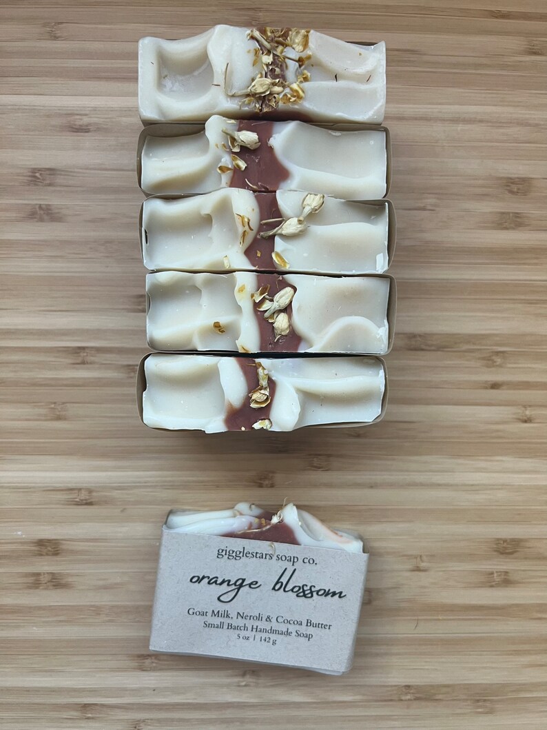 Może przedstawiać: Pięć ręcznie robionych mydeł z białym i brązowym wzorem w wiry, posypanych suszonymi kwiatami. Mydło jest oznaczone "orange blossom" i wykonane z mleka koziego, neroli i masła kakaowego. Każdy kawałek waży 142g.