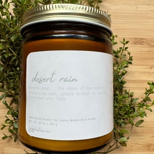 May include: A brown glass candle jar with a white label that reads "desert rain". The label also includes the text "breathe deep. the dance of the desert life giving rain, plants stretch to the sky sun kisses your face". The candle jar is surrounded by green foliage.