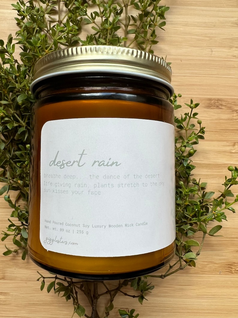 May include: A brown glass candle jar with a white label that reads "desert rain". The label also includes the text "breathe deep. the dance of the desert life giving rain, plants stretch to the sky sun kisses your face". The candle jar is surrounded by green foliage.
