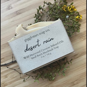 May include: A bar of handmade soap with a paper label that reads "desert rain." The label also states the soap is infused with wild harvested creosote oils and is a small batch product. The soap is 5 oz (142 g). The soap is surrounded by dried greenery on a wooden surface.