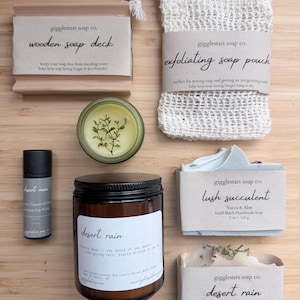 May include: A collection of natural bath and body products, including a wooden soap deck, exfoliating soap pouch, lip balm, a candle, and two soaps. The products are labeled with the brand name "gigglestars soap co." and feature the scent "desert rain."
