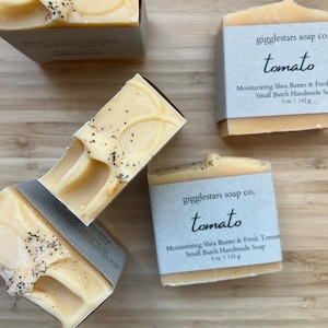 May include: Several bars of handmade soap with the text "tomato" on the label. The soap is light orange with dark flecks and a light-colored design. The label reads "Moisturizing Shea Butter & Fresh Tomato Small Batch Handmade Soap 5 oz | 142 g."