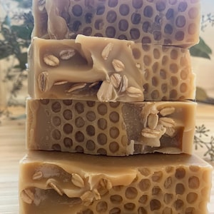 May include: Four bars of honey and oatmeal soap stacked on top of each other. The soap is a light brown color and has a honeycomb pattern. The soap is also speckled with oatmeal.