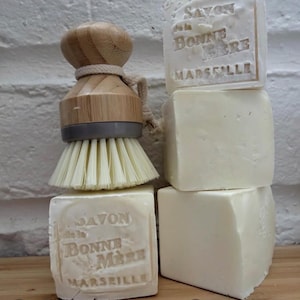 May include: A wooden dish brush with a light-colored bristle head sits atop a stack of four white soap bars. Two bars are stamped with "Savon de la Bonne Mère Marseille". The brush has a wooden handle and a gray base.