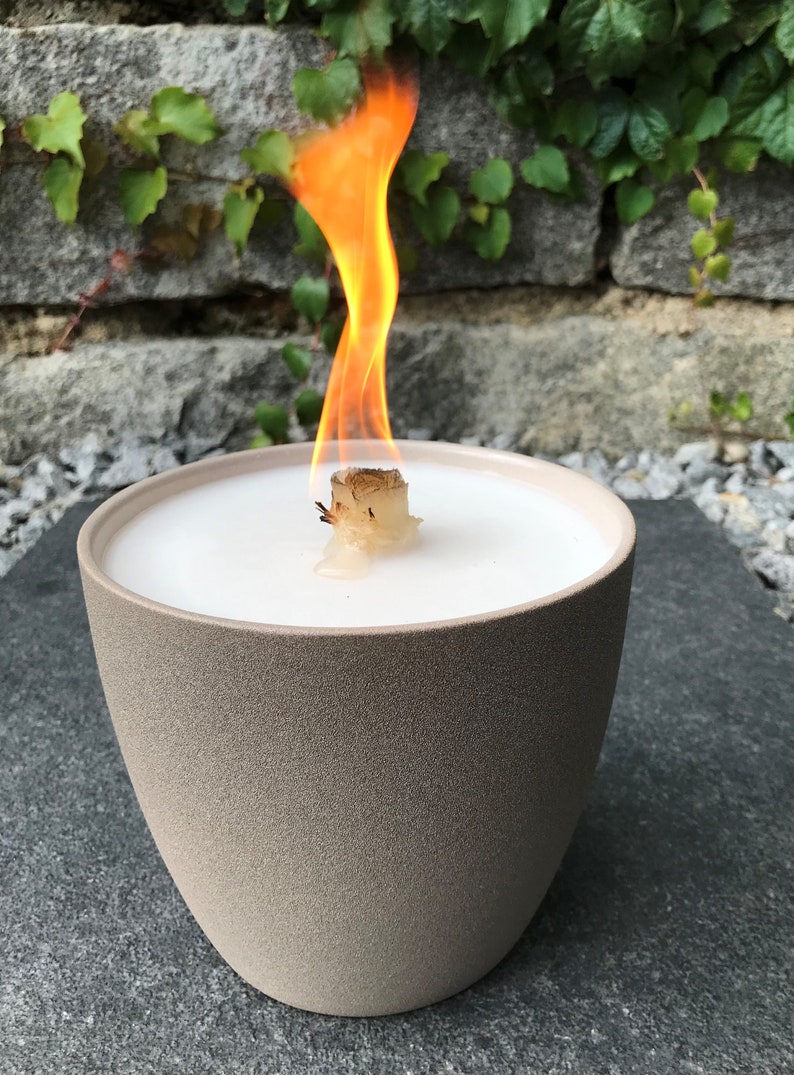 May include: A beige ceramic candle holder with a white wax candle and a wooden wick burning with a bright orange flame.