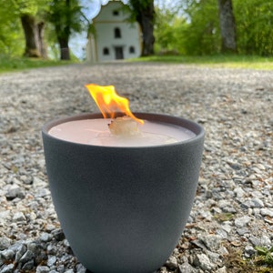 May include: A gray candle holder with a white candle burning inside. The candle is lit and the flame is visible. The candle holder is sitting on a gravel path in front of a white building.