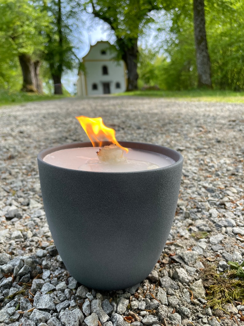 May include: A gray candle holder with a white candle burning inside. The candle is lit and the flame is visible. The candle holder is sitting on a gravel path in front of a white building.