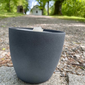 May include: A dark gray ceramic candle holder with a white candle inside. The candle holder is sitting on a gray stone surface.