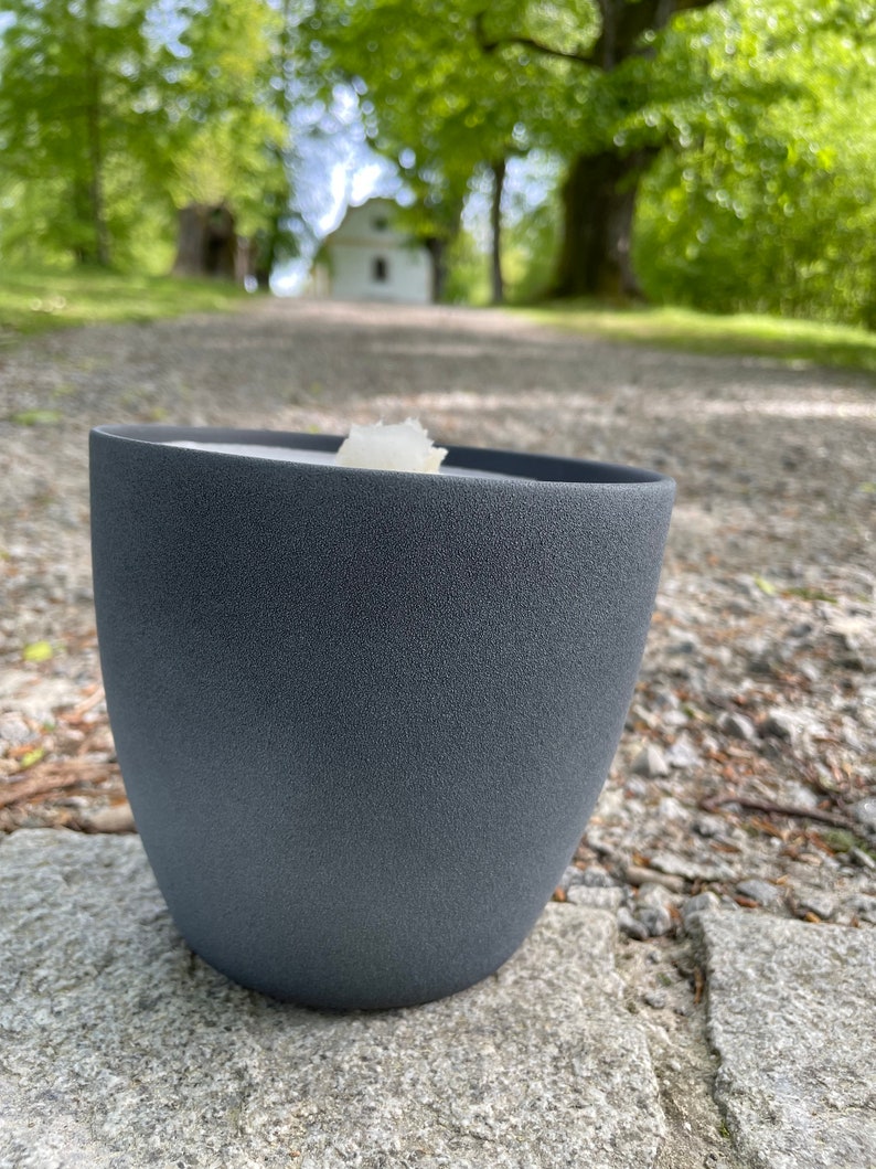 May include: A dark gray ceramic candle holder with a white candle inside. The candle holder is sitting on a gray stone surface.