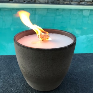 May include: A brown ceramic candle holder with a white candle burning inside. The candle is lit and the flame is visible. The candle holder is sitting on a black surface next to a swimming pool.
