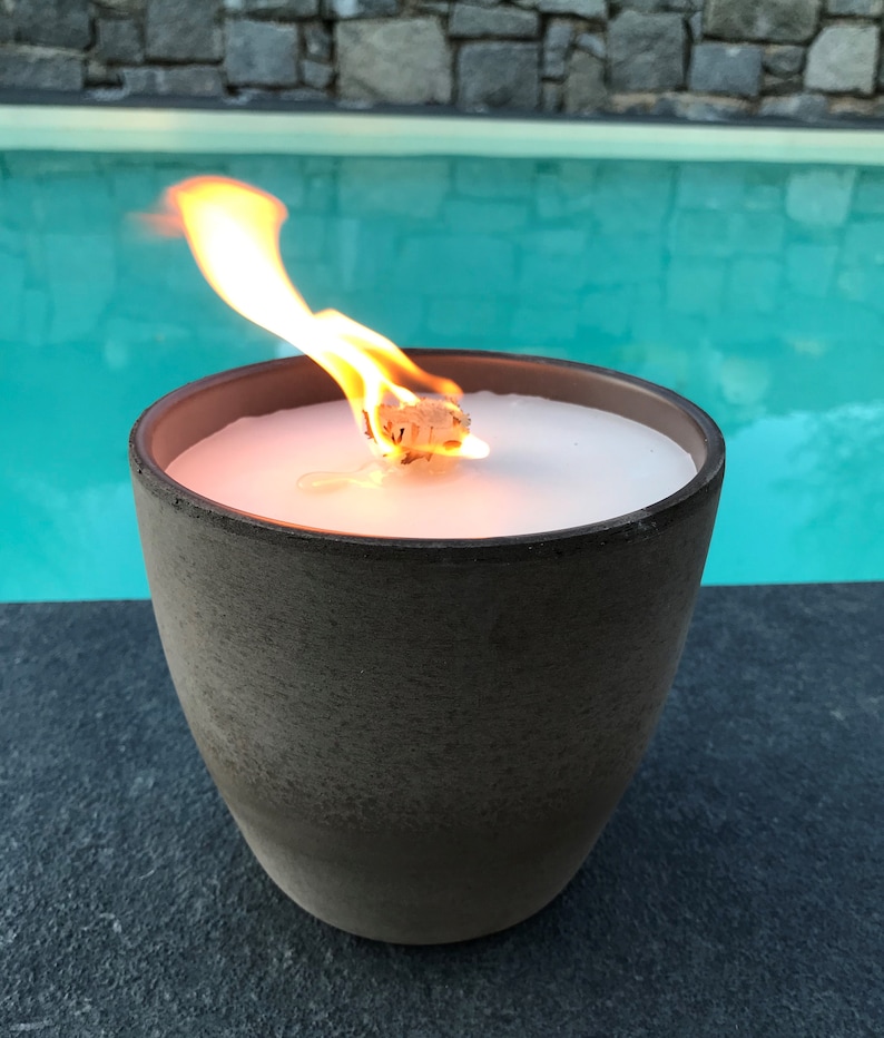 May include: A brown ceramic candle holder with a white candle burning inside. The candle is lit and the flame is visible. The candle holder is sitting on a black surface next to a swimming pool.