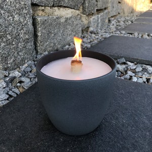May include: A gray ceramic candle holder with a white candle and a wooden wick. The candle is lit and the flame is visible.