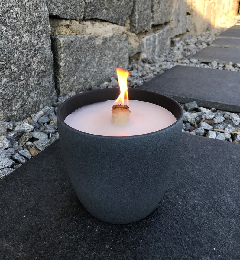 May include: A gray ceramic candle holder with a white candle and a wooden wick. The candle is lit and the flame is visible.