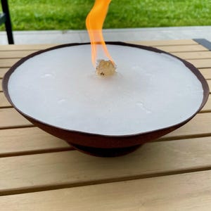 Può includere: Una ciotola per il fuoco rotonda in metallo marrone ruggine con una superficie di combustibile bianca e una fiamma arancione brillante. Il braciere esterno è posto su un tavolo di legno chiaro, con erba verde sullo sfondo.