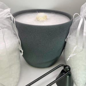Op de afbeelding: Een grijze keramische kaarsenpot met een witte waskaars erin. Twee witte gaaszakjes gevuld met witte badzout bevinden zich aan weerszijden van de kaarsenpot. Een zwarte metalen kaarsdover bevindt zich op de voorgrond.