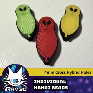 May include: Three colorful alien-shaped Kandi beads. The beads are green, red, and yellow, each with a black outline and a smiling face. The text "4mm Cross Hybrid Holes" and "INDIVIDUAL KANDI BEADS" are also visible.