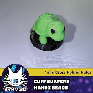 May include: A green plastic turtle charm with black eyes, sitting on a black base with holes. The base is labeled "4mm Cross Hybrid Holes" and "CUFF SURFERS KANDI BEADS".