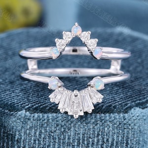 May include: A silver ring with a unique design. The ring features a double band, with a top band adorned with small white gemstones and opals, and a lower band with a fan-shaped arrangement of baguette-cut stones. The ring is set against a blue background.