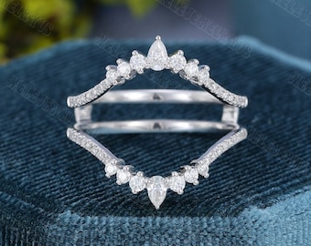 May include: A silver ring guard with a double band design. The ring features a series of small, round, and pear-shaped clear gemstones. The ring is set against a dark teal background.