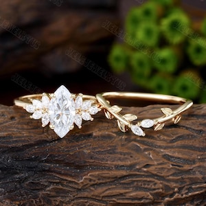 May include: A gold engagement ring set featuring a marquise-cut center stone surrounded by smaller stones, and a matching band with leaf and stone accents. The rings are displayed on a dark wooden surface.