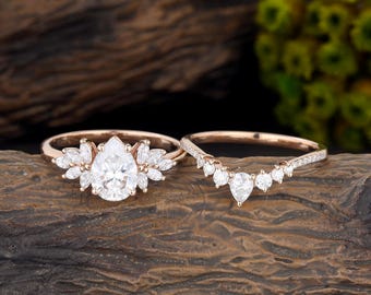 May include: A rose gold engagement ring set featuring a central pear-cut stone surrounded by marquise-cut side stones, paired with a curved wedding band adorned with a series of small round-cut stones along the band and a pear-cut stone.