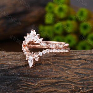May include: A rose gold ring with a delicate, curved design featuring a row of small diamonds and a larger marquise-shaped diamond at the center. The ring is set on a wooden surface.