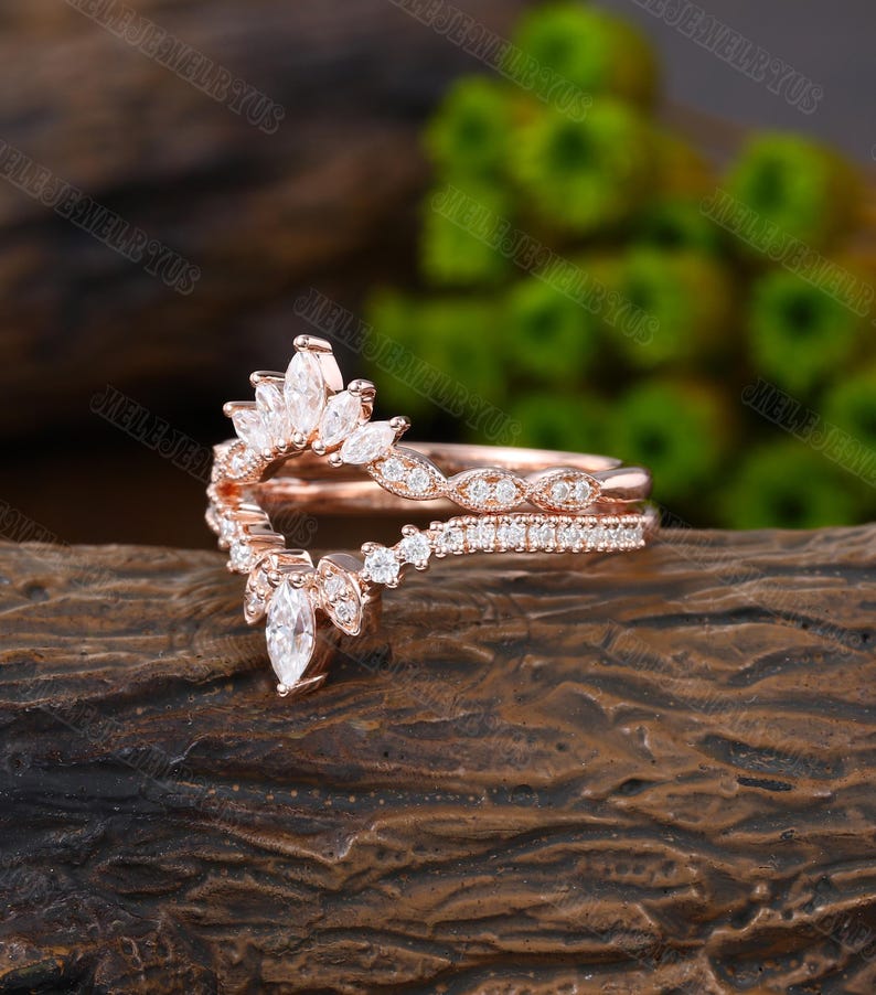 May include: A rose gold ring with a delicate, curved design featuring a row of small diamonds and a larger marquise-shaped diamond at the center. The ring is set on a wooden surface.